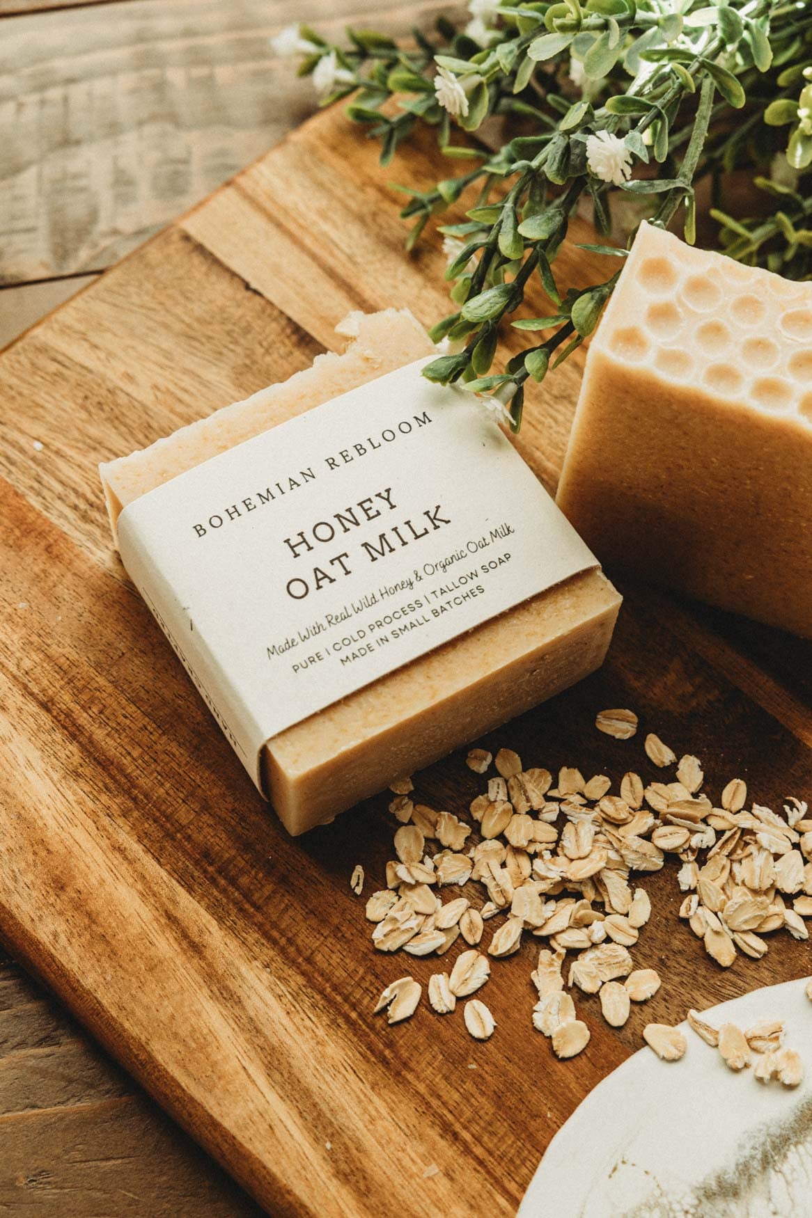 Bar of soap labeled 'Honey Oat Milk' on a wooden board with oats and greenery.
