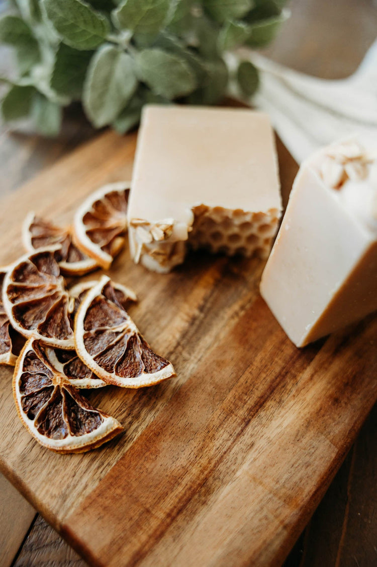 Two pieces of honey comb oat milk soap with slices of dried lemon on a wooden cutting board.