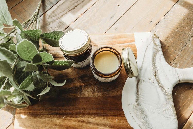 Two jars of herbal tallow on top of wooden cutting with greenary in the background.