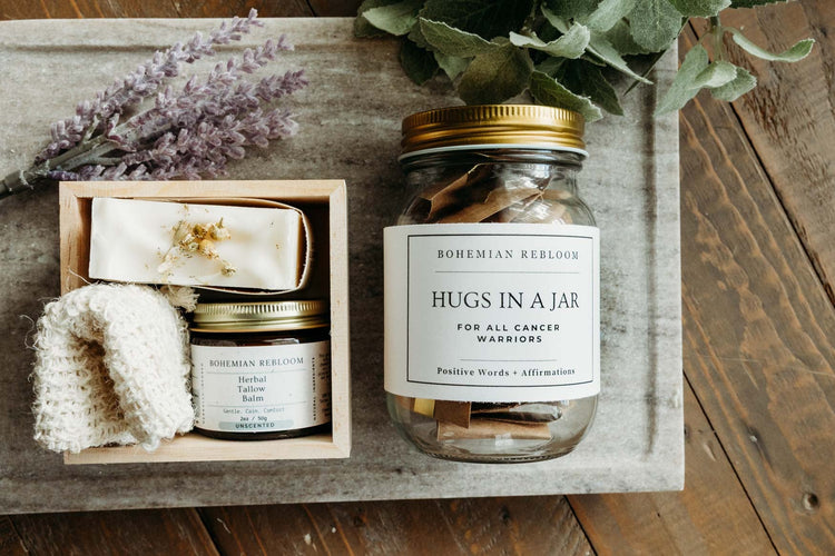 A glass mason jar filled with inspirational cardstock messages and a gift box of chamomile lavender tallow soap, soap saver, and herbal tallow balm in a gift set on top of a marble top with flowers and greenery in the background.