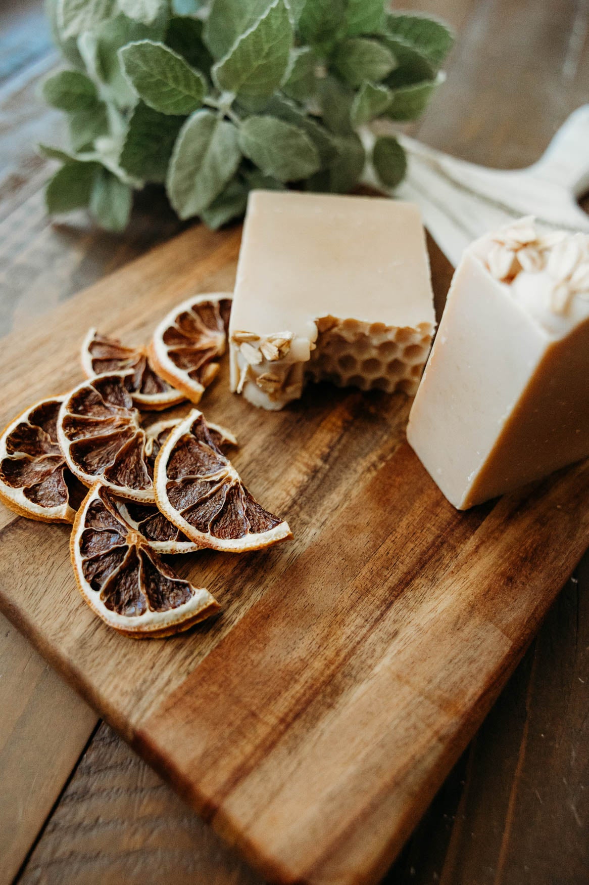 Two blocks of honey oat milk 
soap on a wooden cutting board with dried lemon slices and a plant in the background.