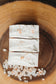 Three square pieces of pink himalayan sea salt soap with himalayan sea salt crystals on top a wooden surface.