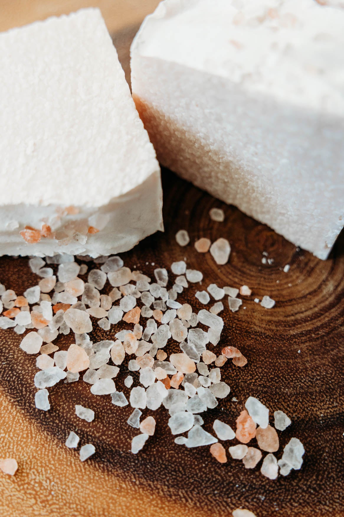 Two pieces of himalayan sea salt soap on a wooden surface with small bath salts.