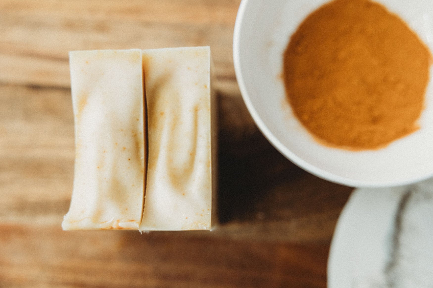 Two bars of honey chai turmeric soap next to each other with a bowl of turmeric powder on a wooden surface