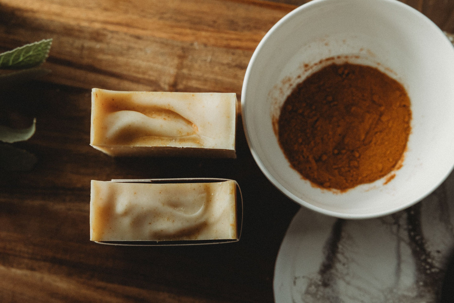Two bars of honey chai turmeric soap with a bowl of turmeric powder on a wooden surface