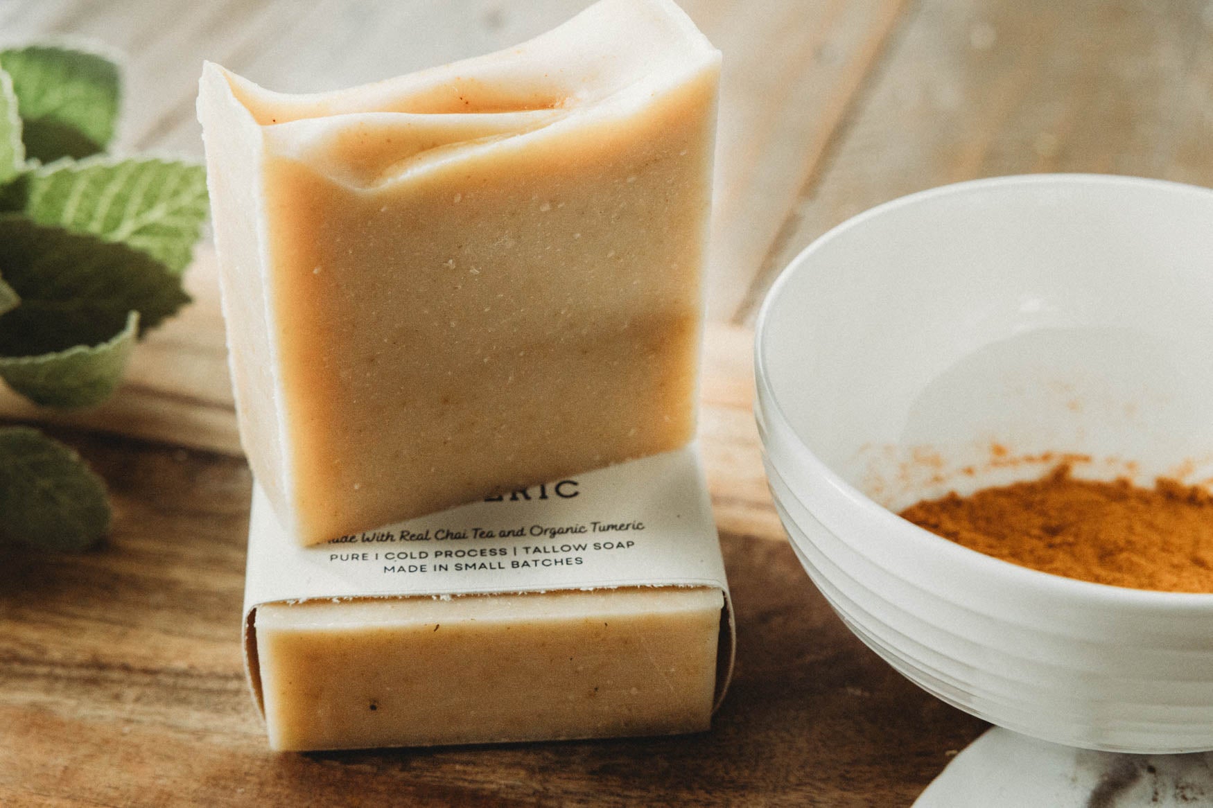 Chai turmeric honey bar of soap with a visible brand label on a wooden surface, next to a white bowl and green leaves.