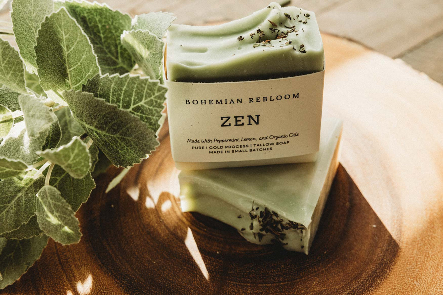 Two bars of green soap with 'Bohemian Rebloom' packaging on a wooden surface with green leaves.