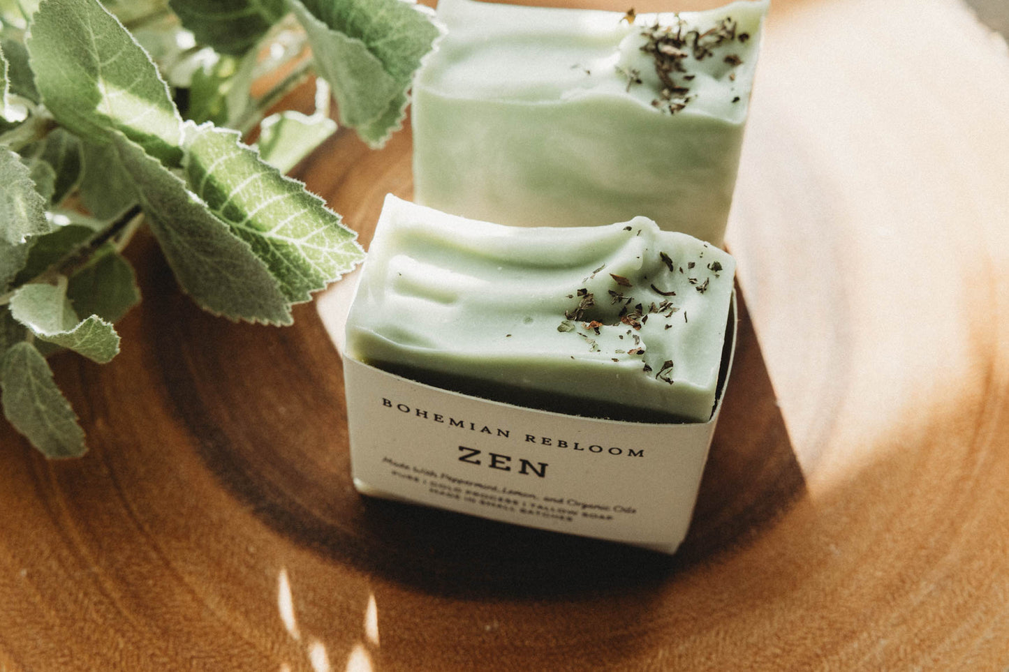 Two bars of green and white swirled soap labeled 'Zen' on a wooden surface with greenery.