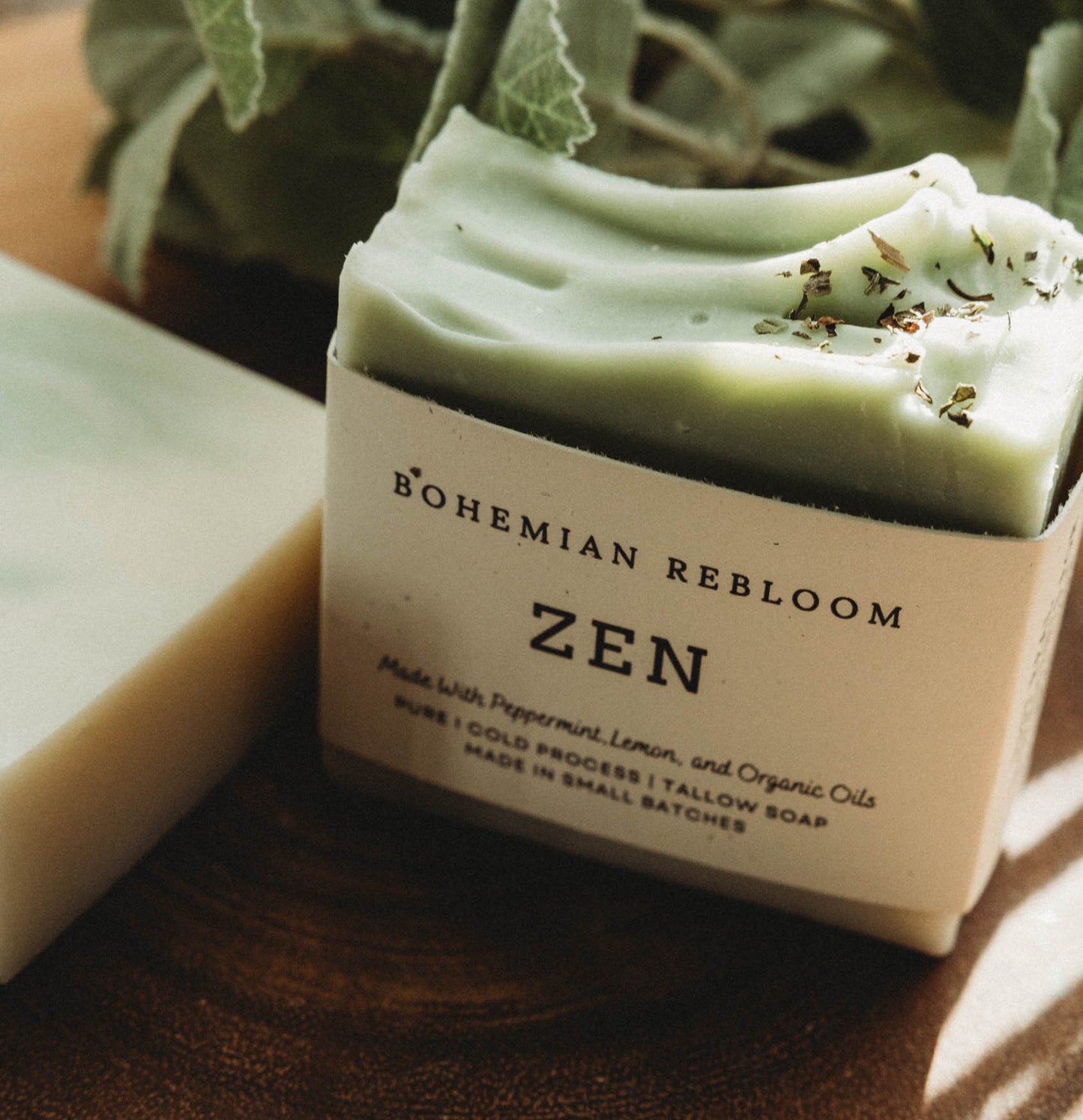 Green bar of cold process soap with 'Bohemian Rebloom Zen' label on a wooden surface with greenary in the background.
