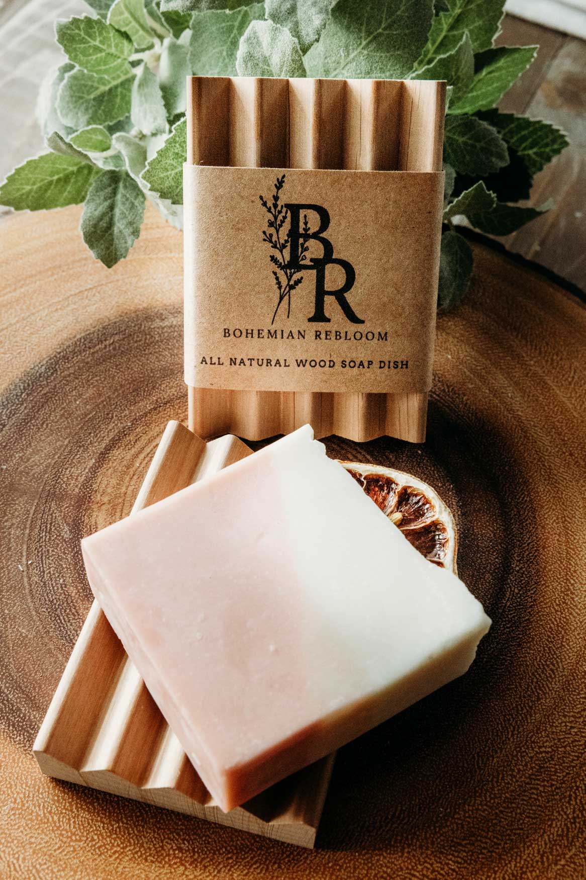 A Pink Lemonade bar of soap with the Bohemian Rebloom label and another bar of soap on top of a wooden soap dish with a plant in the background