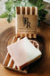 A Pink Lemonade bar of soap with the Bohemian Rebloom label and another bar of soap on top of a wooden soap dish with a plant in the background