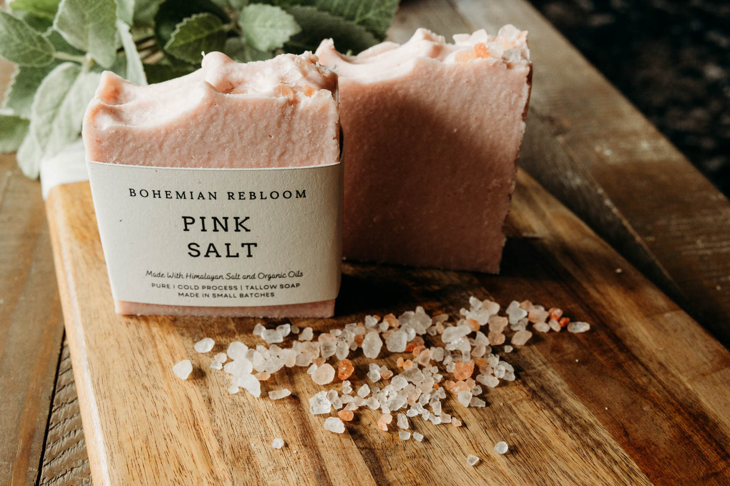 Two bars of pink salt soap with a label on a wooden board, surrounded by sea salt crystals.