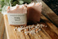 Two bars of pink salt soap with a label on a wooden board, surrounded by sea salt crystals.