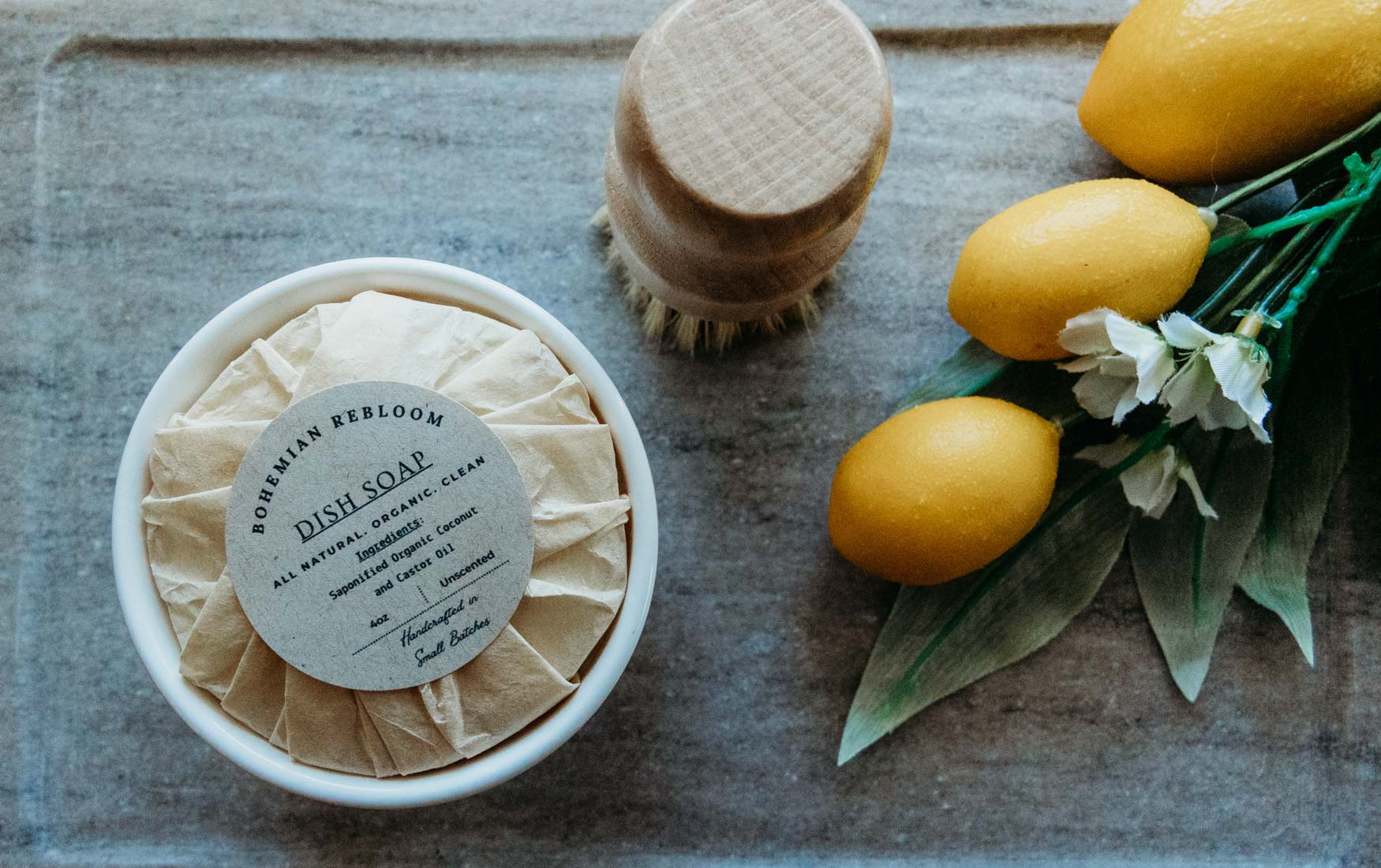 Ramekin dish with Bohemian Rebloom label filled with organic coconut and castor oil dish soap with brush on top on a marble board with lemons.