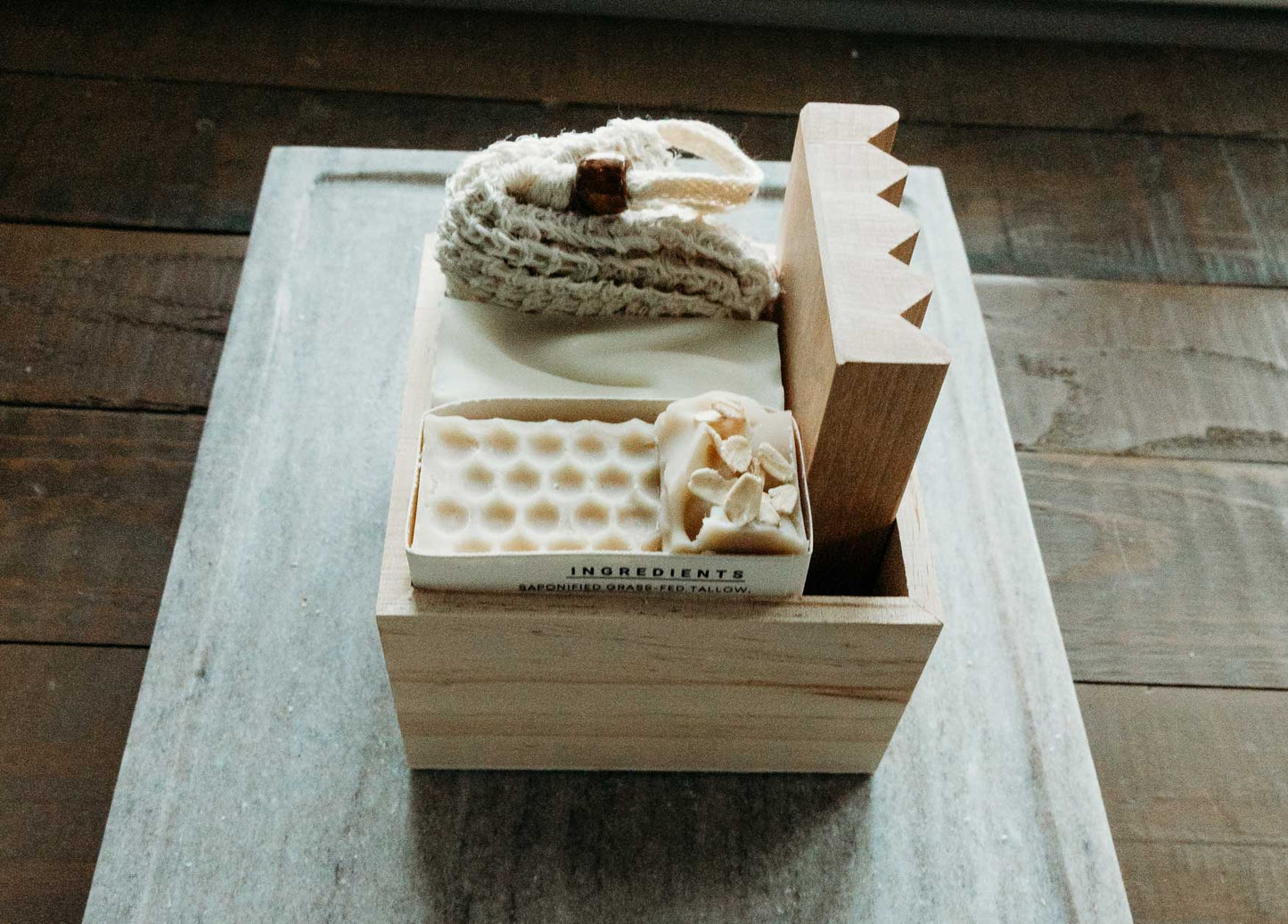 Wooden box with a bar of sweet almond and honey oat milk soap along with a soap saver bag and wooden dish holder on top of a marble top.