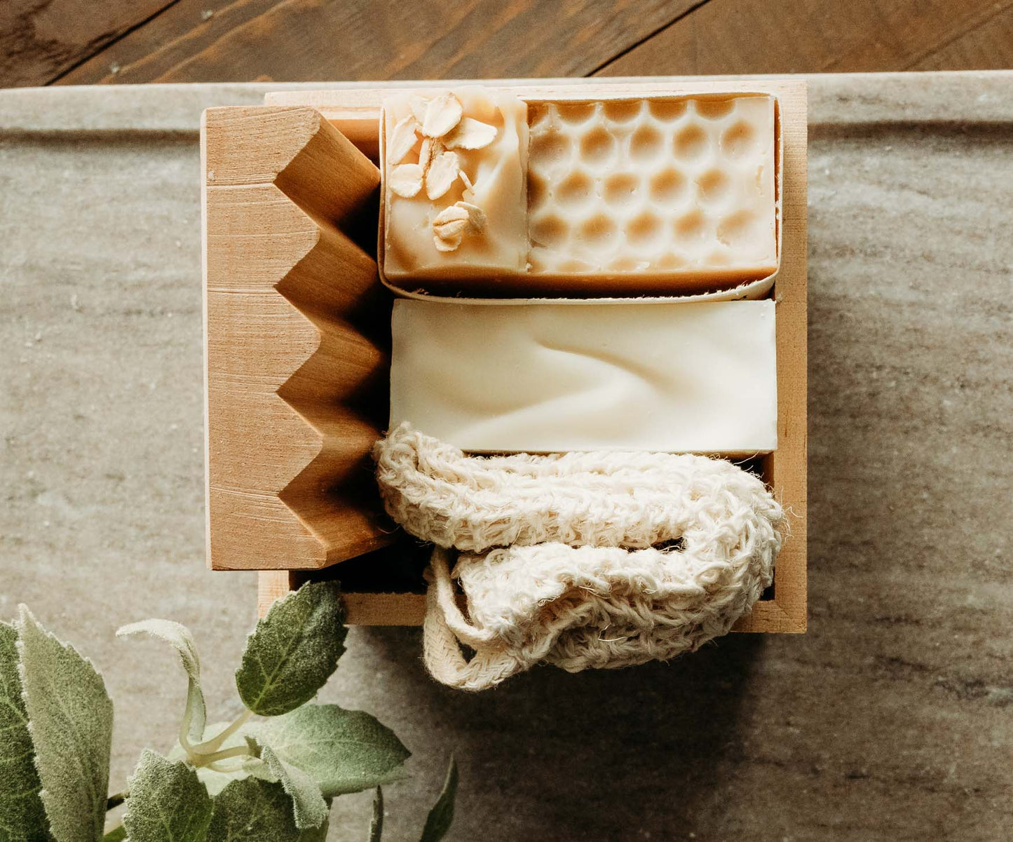 A bar of sweet almond and honey oat milk soap with a soap saver and  wooden soap dish on a marble table with greenery in the background.