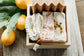 A bar of himalayan sea salt soap, lavender chamomile soap, a soap saver, and wooden soap dish onto of a marble board with lemons in the background.