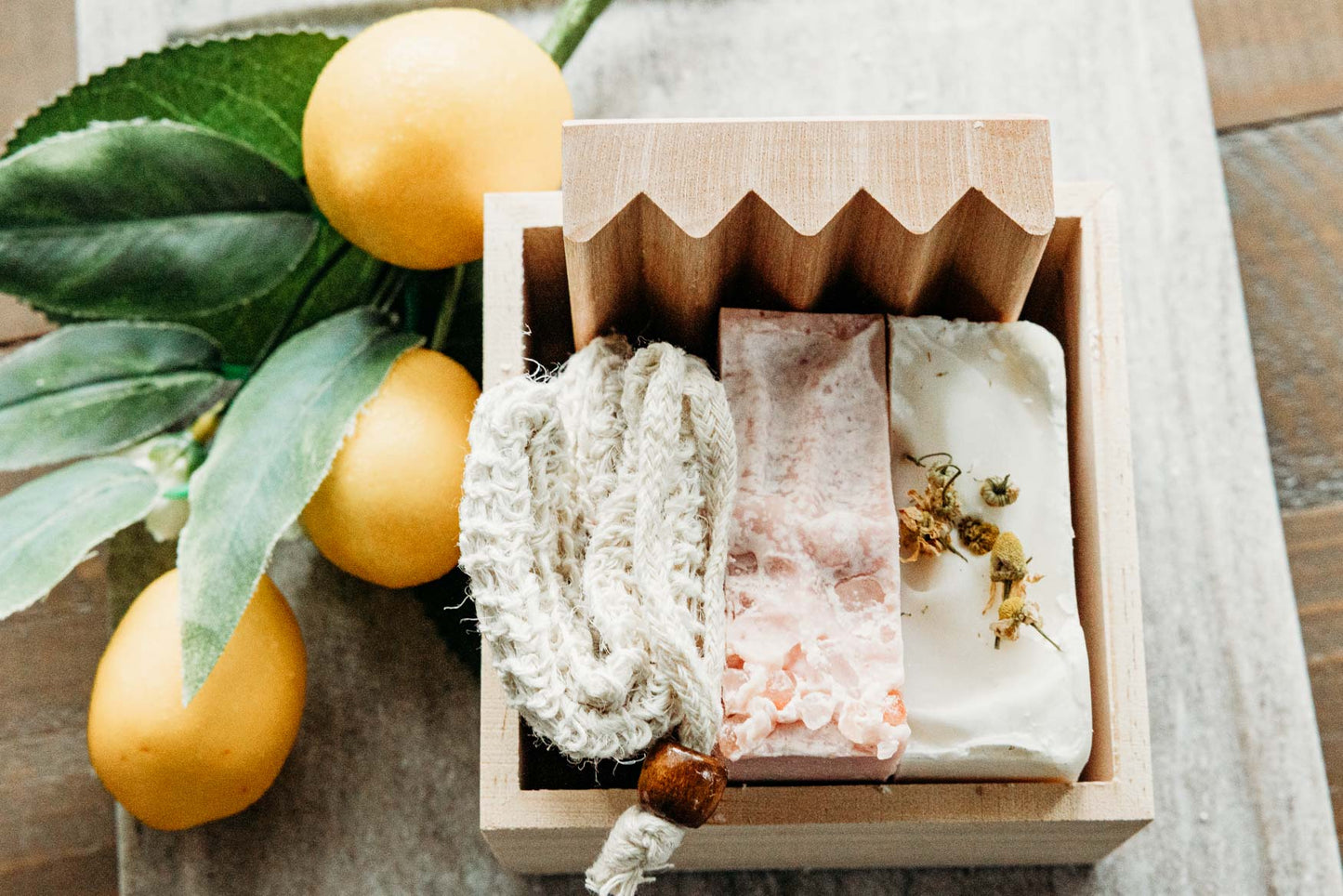 A bar of himalayan sea salt soap, lavender chamomile soap, a soap saver, and wooden soap dish tucked into a wooden box onto of a marble board with lemons in the background.