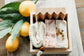 A bar of himalayan sea salt soap, lavender chamomile soap, a soap saver, and wooden soap dish tucked into a wooden box onto of a marble board with lemons in the background.