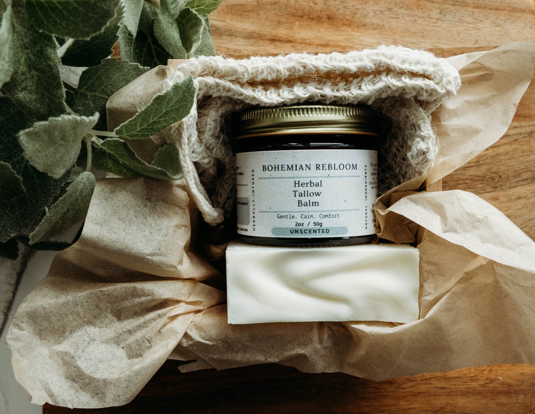 Jar of Bohemian REbloom Herbal Tallow Balm, Sweet Almond Soap, and soap saver  in a box on top of a wooden surface with greenery.