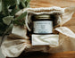 Jar of Bohemian REbloom Herbal Tallow Balm, Sweet Almond Soap, and soap saver  in a box on top of a wooden surface with greenery.
