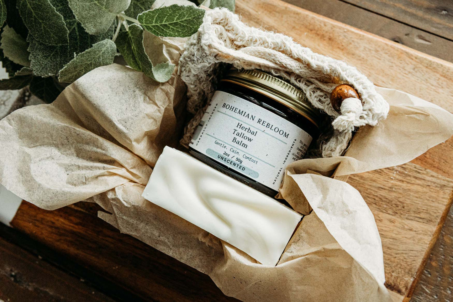 Side view of a jar of Bohemian REbloom Herbal Tallow Balm, Sweet Almond Soap, and soap saver  in a box on top of a wooden surface with greenery.
