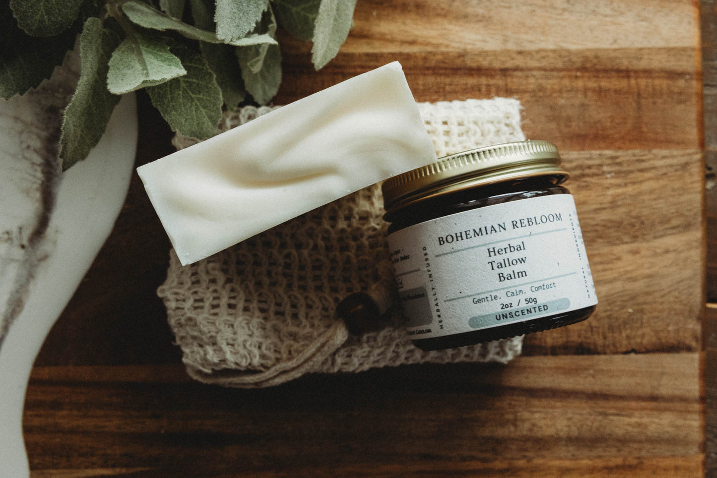 A jar of Bohemian Rebloom Herbal Tallow Balm, Sweet Almond Soap, and soap saver  bag on top of a wooden surface with greenery.