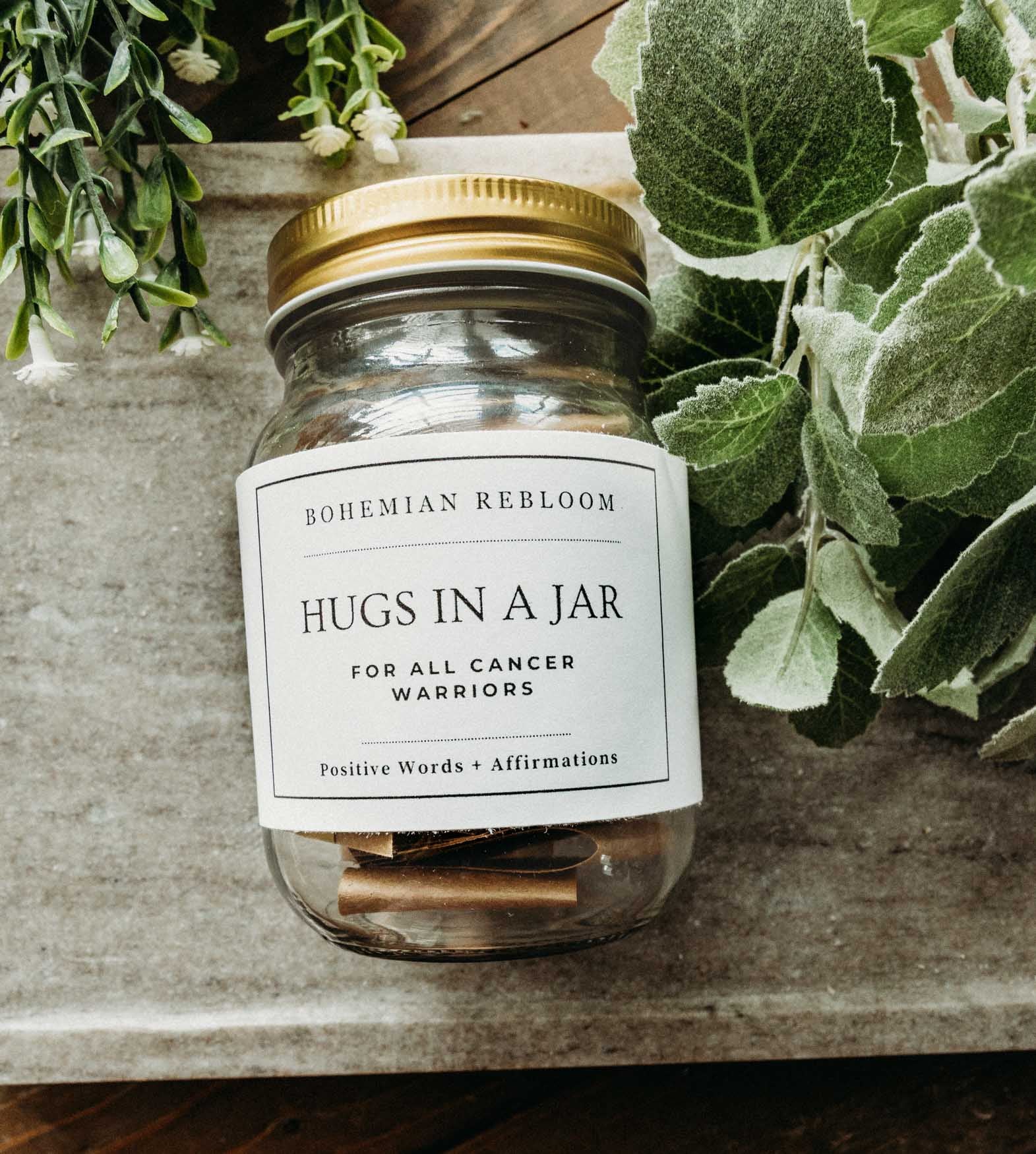 Jar labeled 'Hugs in a Jar' with a gold lid on a wooden surface surrounded by greenery.