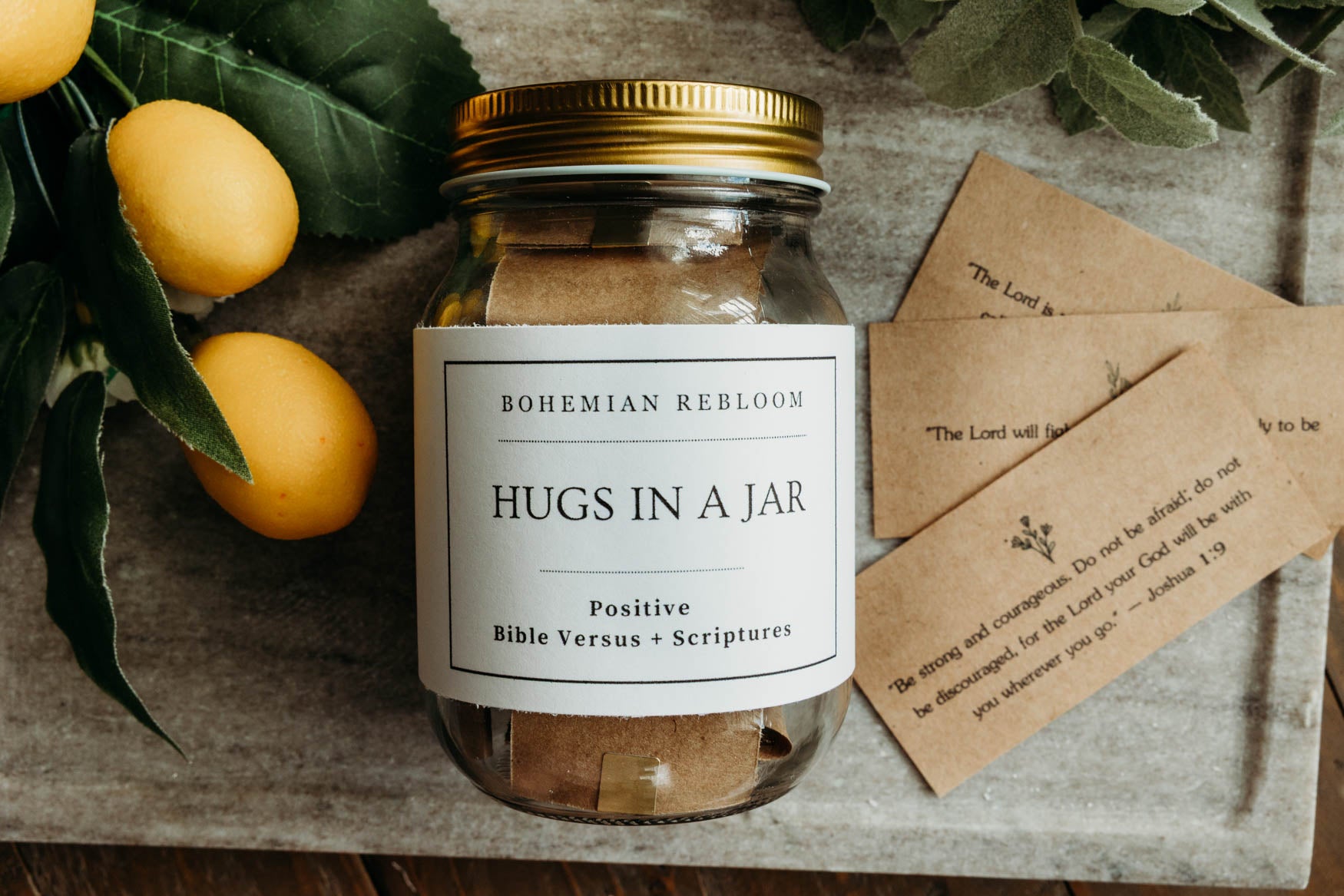 Jar labeled 'Hugs in a Jar' filled with positive Bible versus and scriptures  and cards on a wooden surface with lemons in the background.