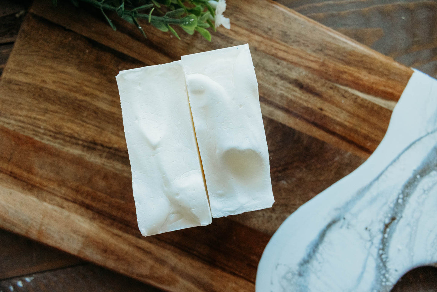 Two pieces of grass fed tallow soap on a wooden cutting board with greenery in the background.