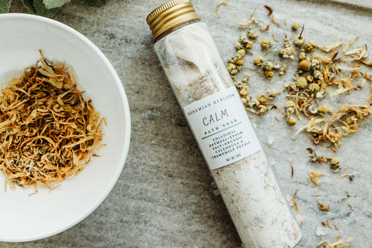 Bath salt tube labeled 'Calm' next to a bowl of dried herbs on a textured surface