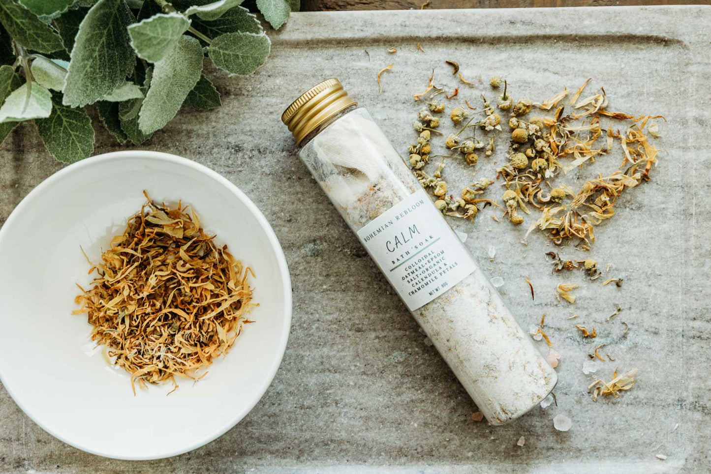 Bottle of bath salts labeled 'Calm' on a stone surface with dried herbs and a leafy plant.