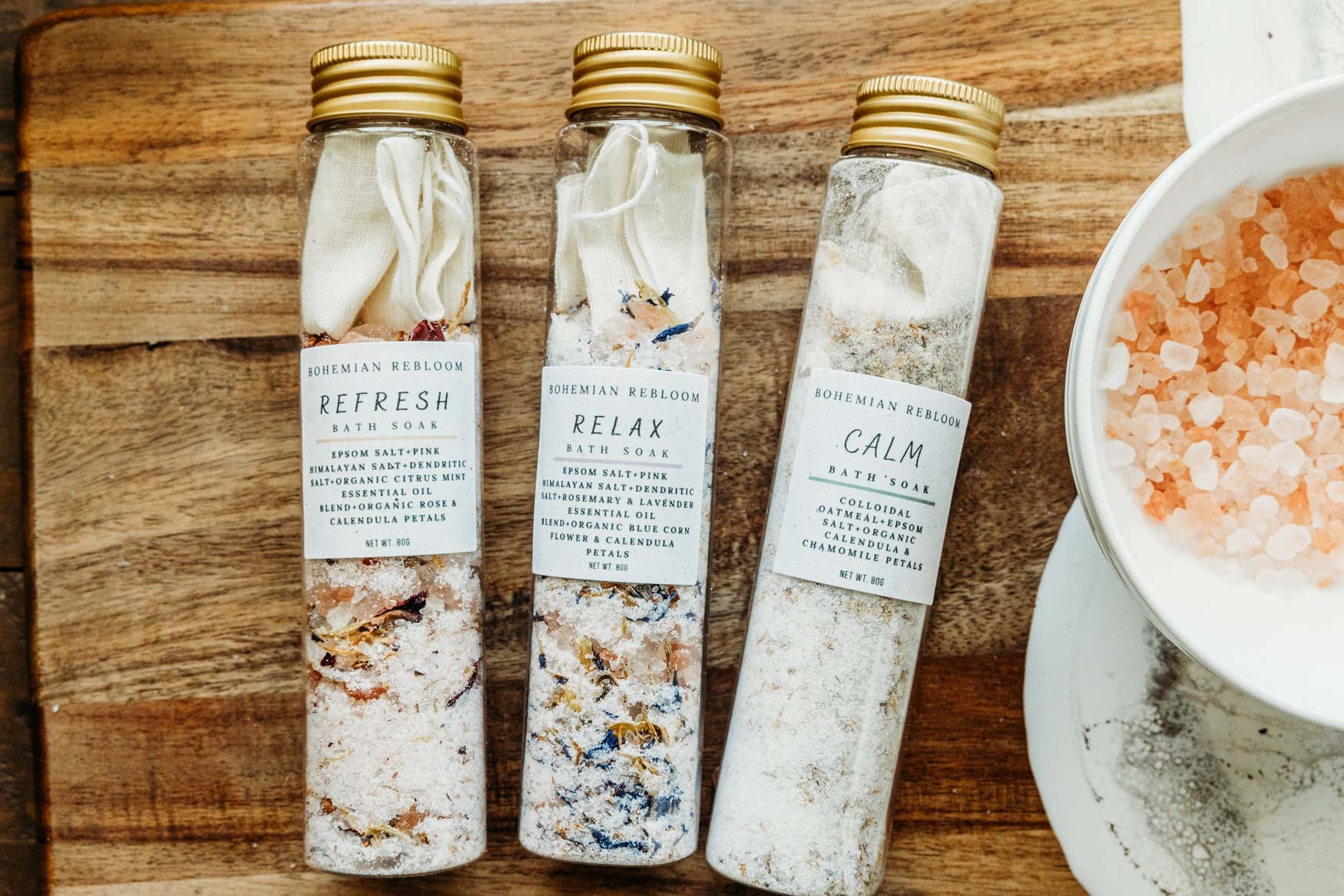 Three test tube bottles of bath salts laying flat on a wooden cutting board with a bowl of salts nearby.