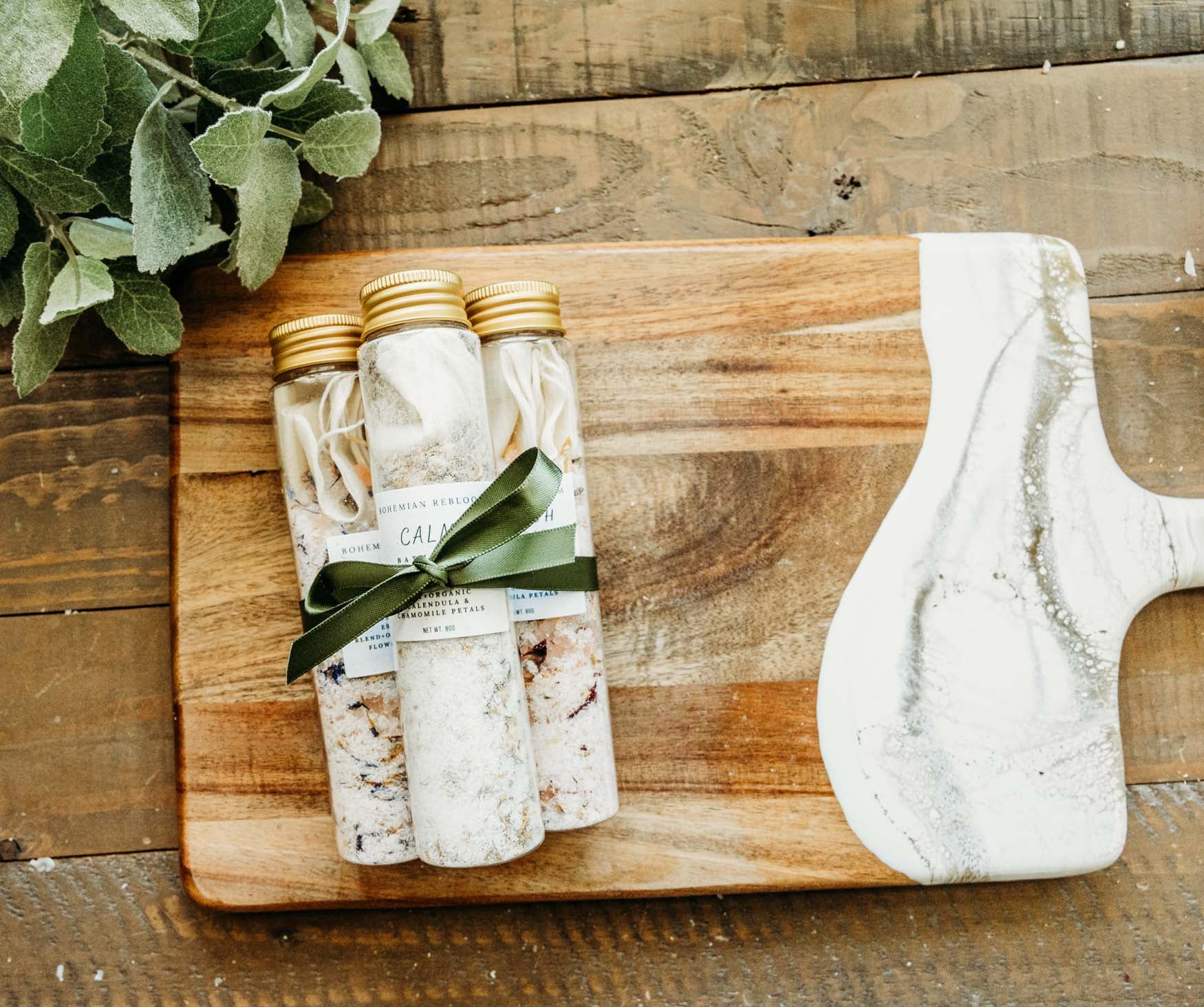 Three bottles of bath salts wrapped  with a green bow on a wooden cutting board.