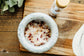 Bowl of bath salts on a wooden surface with a bottle and plant in the background