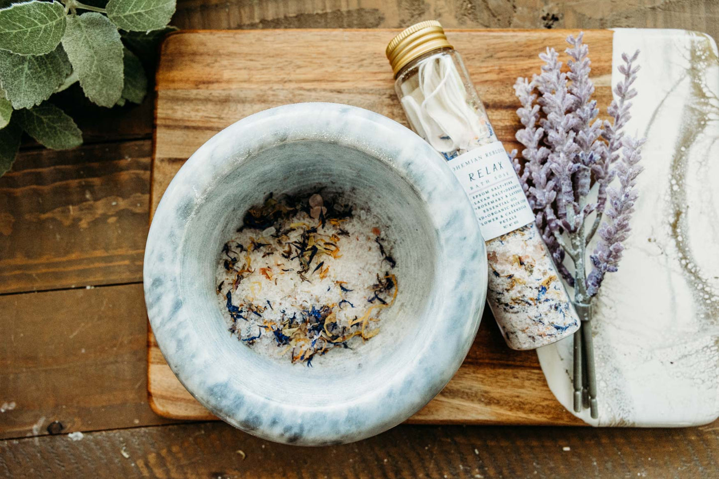 Mortar and pestle with dried herbs on a wooden board, next to a bottle of bath salts and lavender flowers.