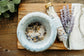 Mortar and pestle with dried herbs on a wooden board, next to a bottle of bath salts and lavender flowers.