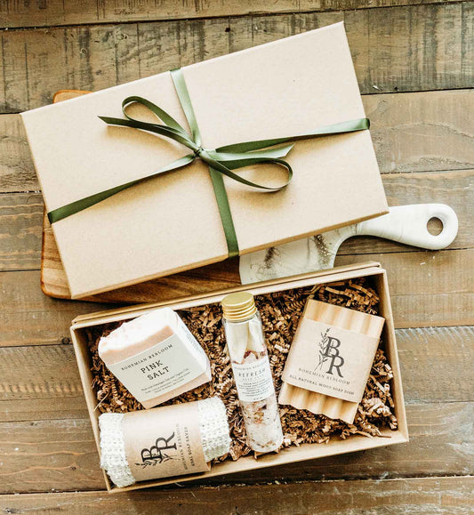 Gift set with a pink Himalayan salt soap bar, refresh bath salts, sisal soap saver, and wooden soap dish in a box on top of a wooden surface. 