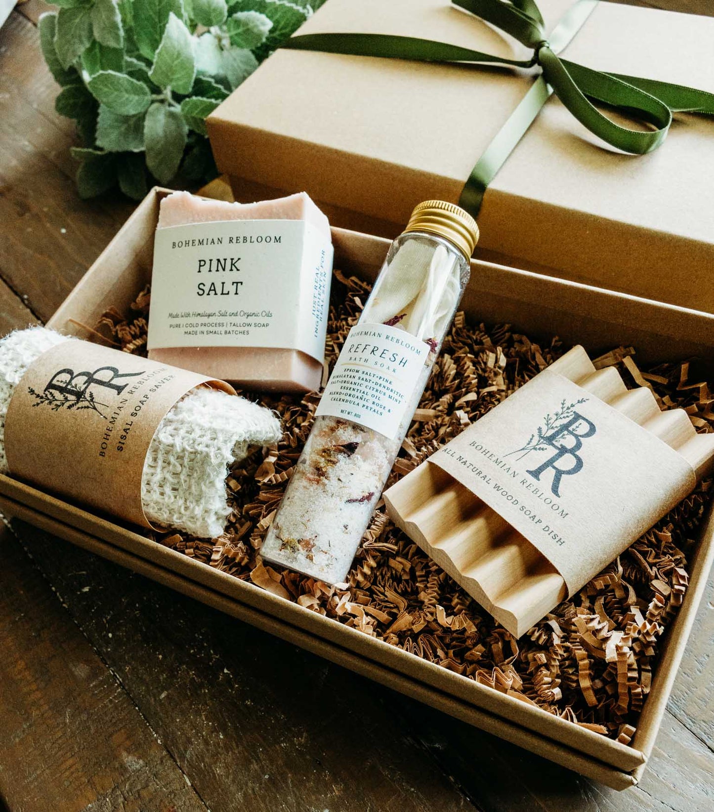 Gift set with bath products including soap, bath salt test tube, sisal soap saver bag, and wooden soap dish in a box on a wooden surface.