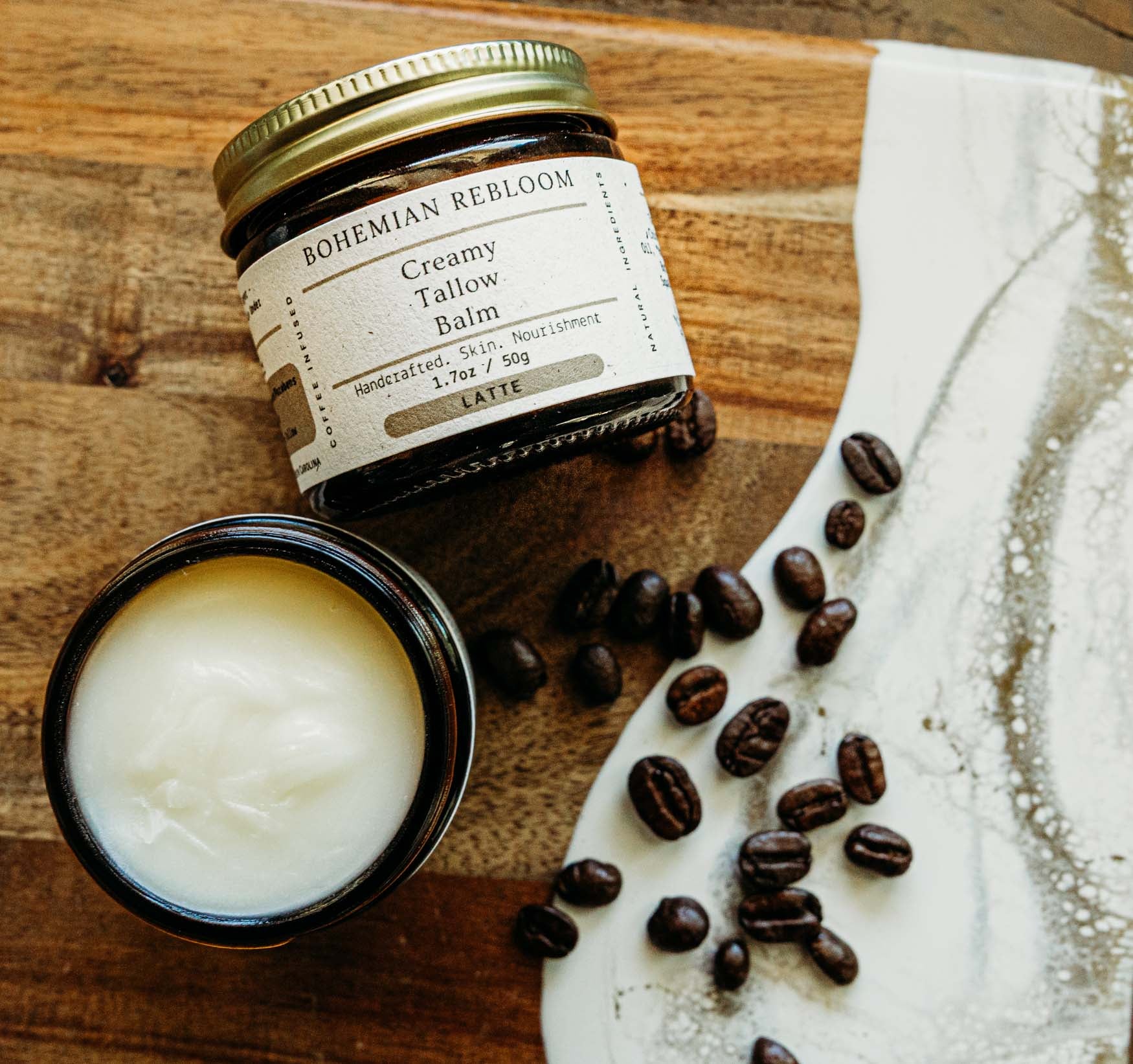 Jar of Bohemian ReBloom Creamy Tallow Balm on a wooden surface with coffee beans and a white cloth.