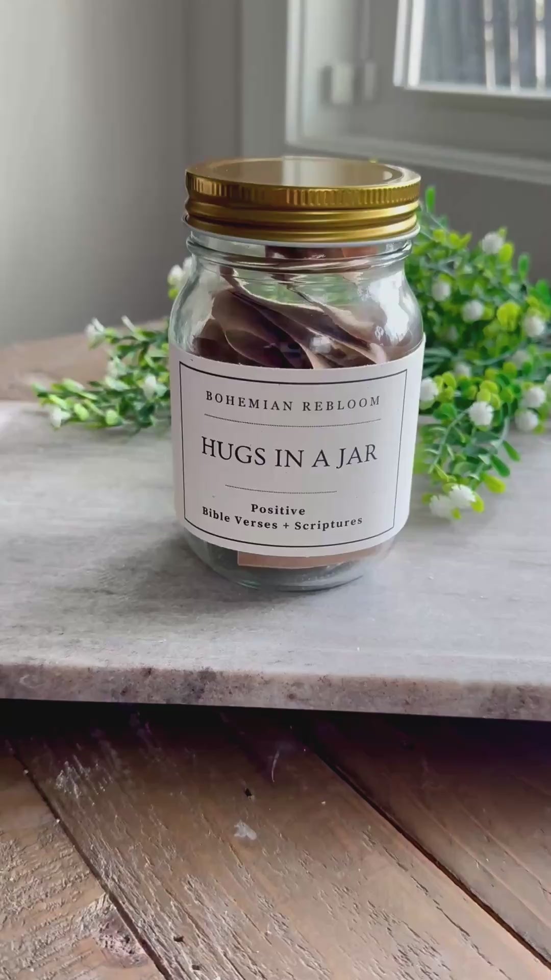 A glass mason jar with a gold lid with a label that has the Bohemian Rebloom brand name and states Hugs In A Jar - Positive Bible verses and scriptures. The back of the jar is then shown as well as the opening of one of the positive verse cardstock notecards inside the mason jar. 